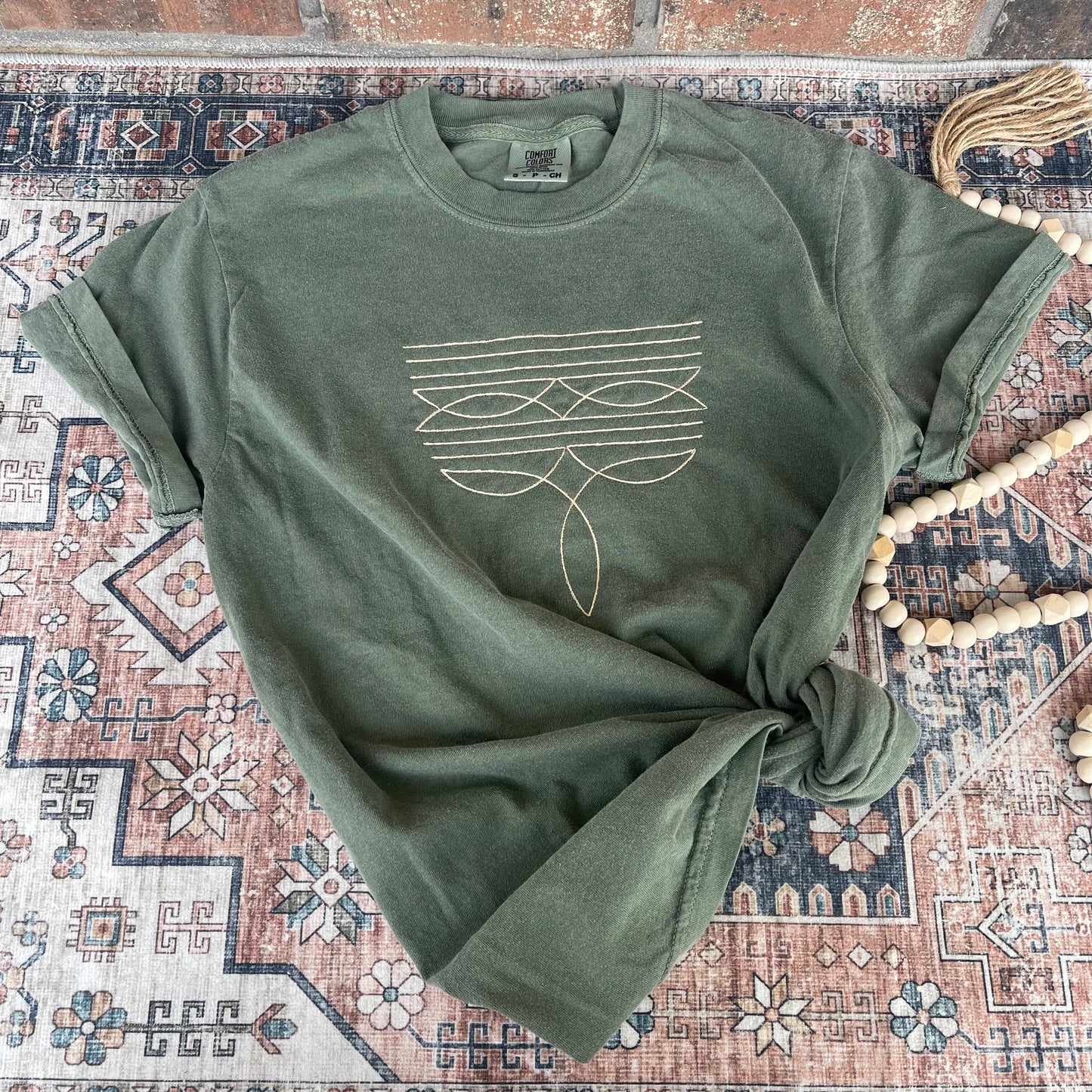 A green Comfort Colors tee with unique boot stitching detail laid out on a patterned surface.