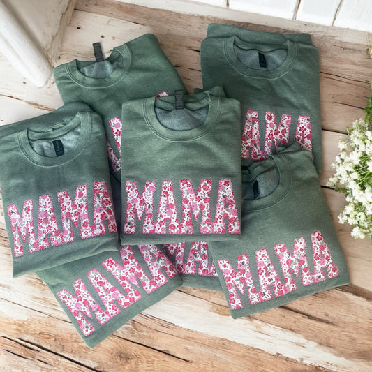 Green sweatshirts with 'MAMA' printed in pink floral letters on a wooden surface.