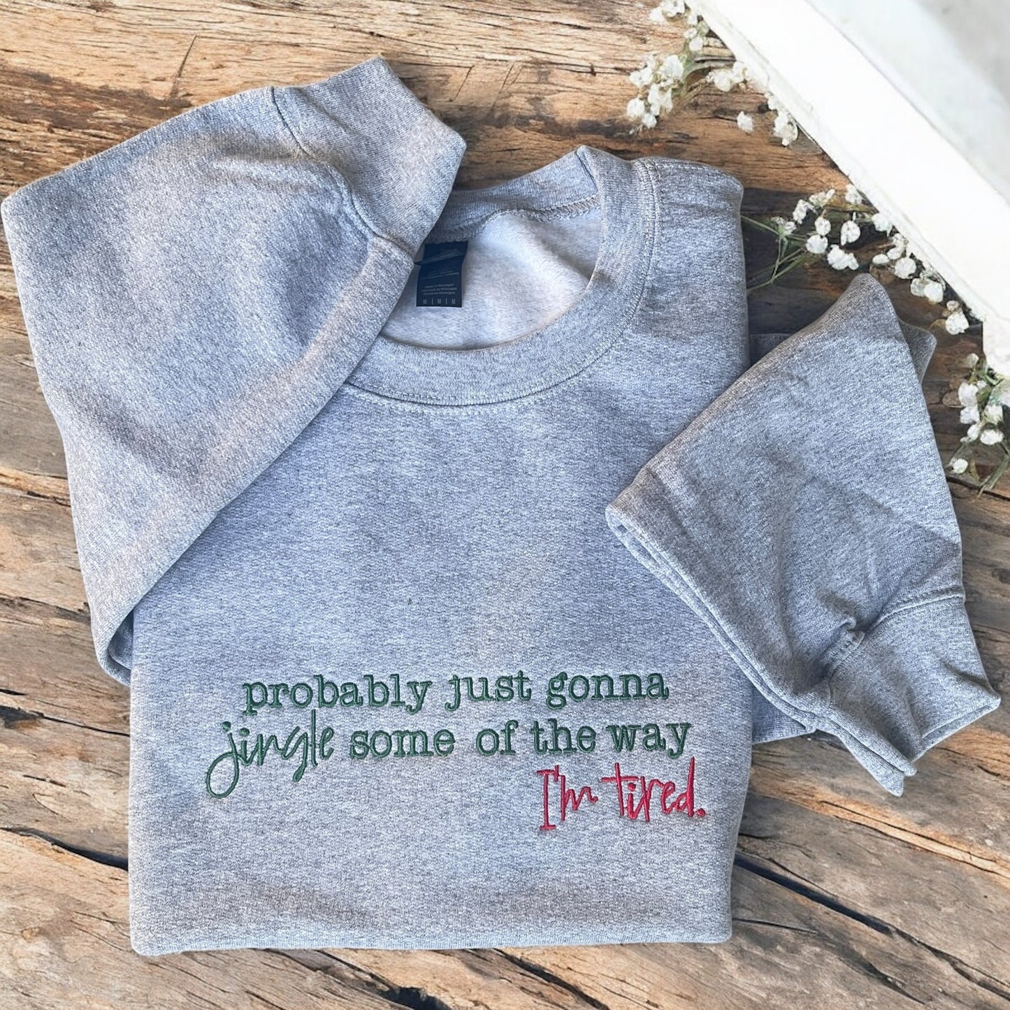 Folded gray sweatshirt with humorous text on a wooden surface with flowers.