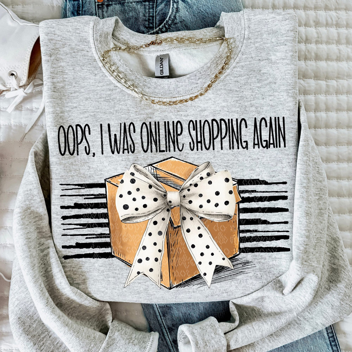 Gray sweatshirt with a graphic of a box with a bow and text 'Oops, I was online shopping again' on a textured surface.