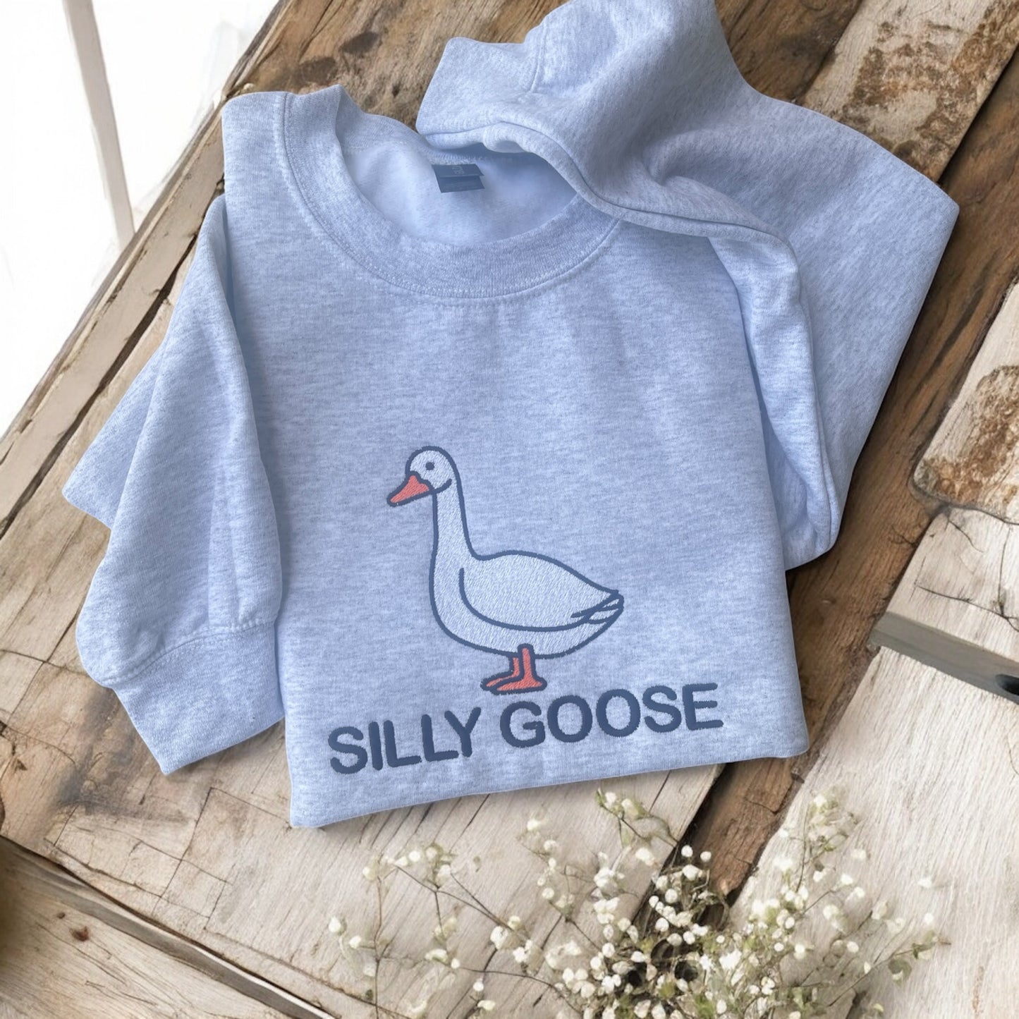 Light blue sweatshirt with a goose graphic and 'Silly Goose' text on a wooden surface.