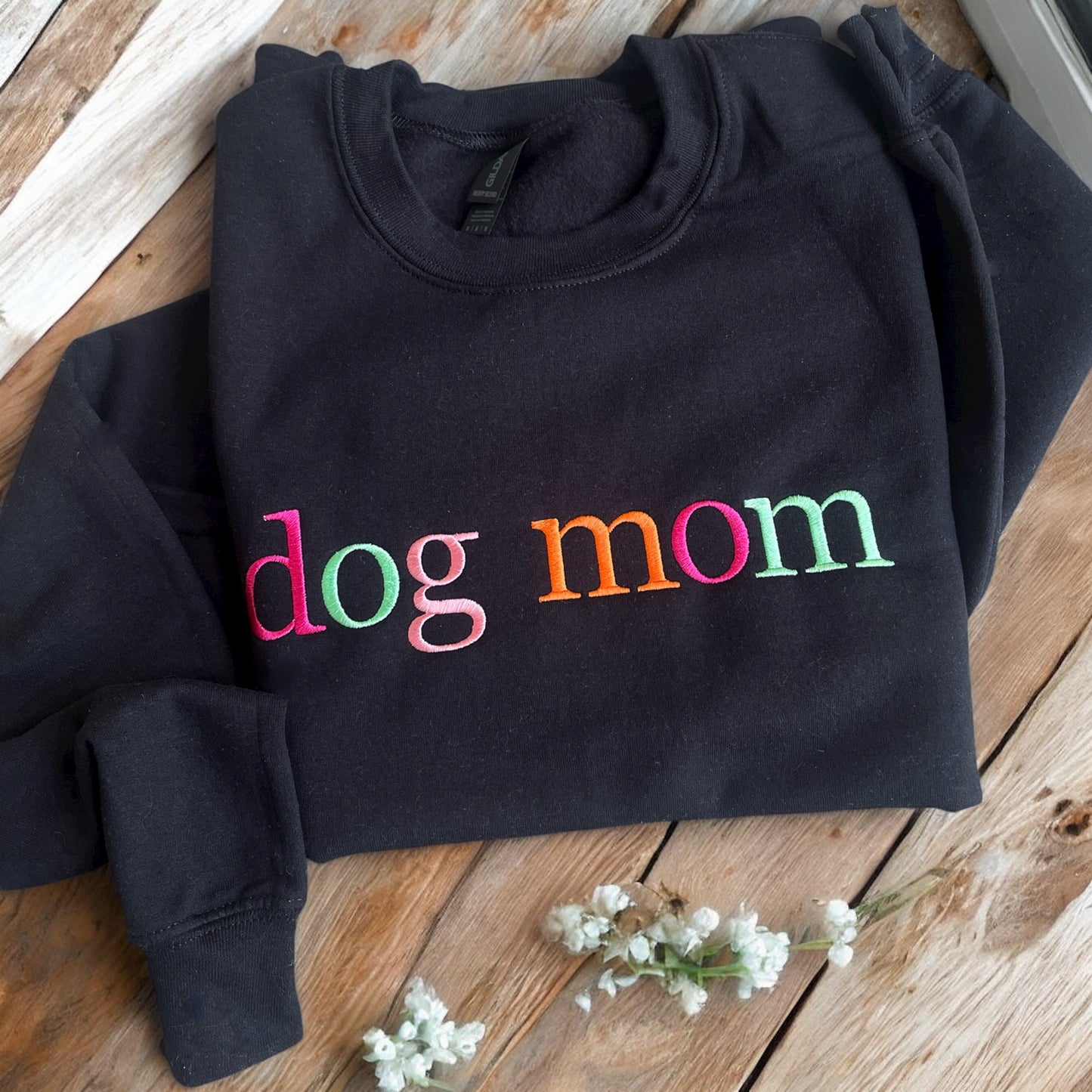 Black hoodie with 'dog mom' in colorful letters on a wooden surface with white flowers.