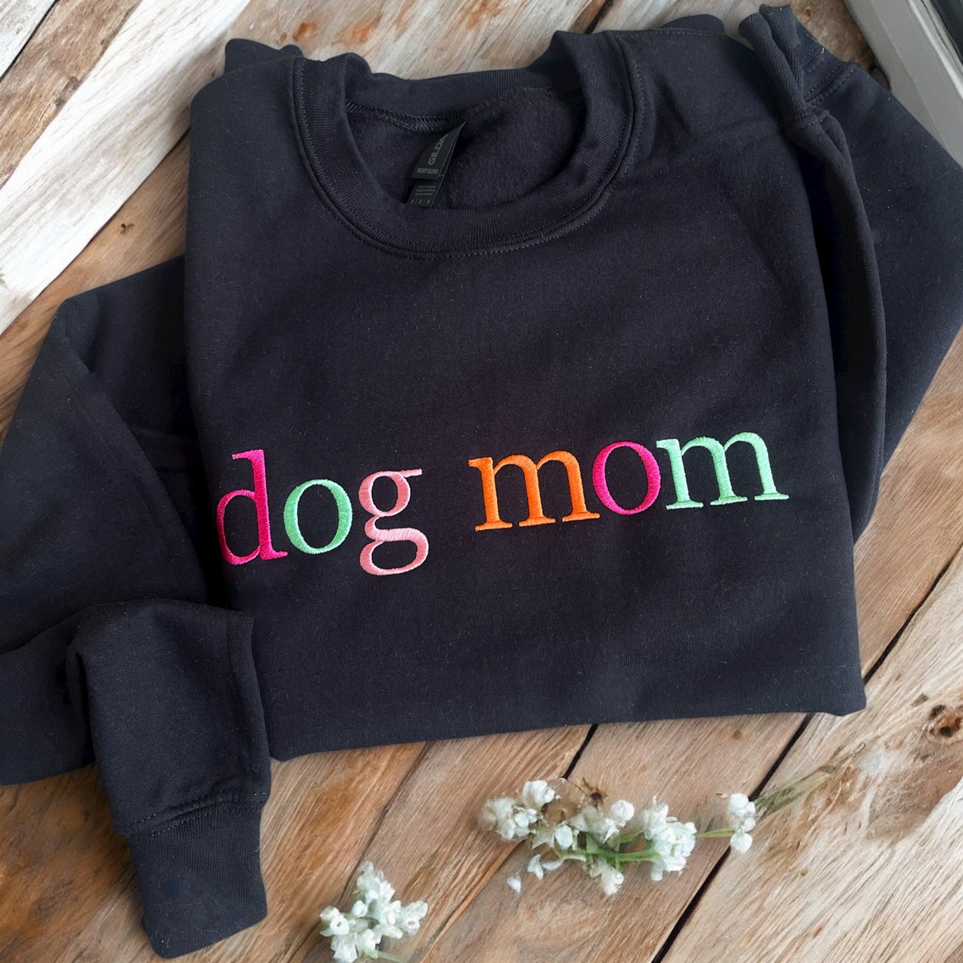 Black hoodie with 'dog mom' in colorful letters on a wooden surface with white flowers.
