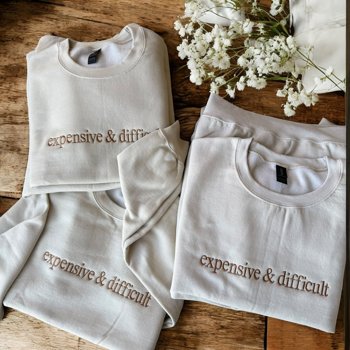 White sweatshirts with 'expensive & difficult' text on a wooden surface with white flowers.