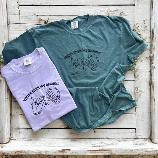 Two t-shirts, one green and one purple, with a graphic design on a wooden surface.