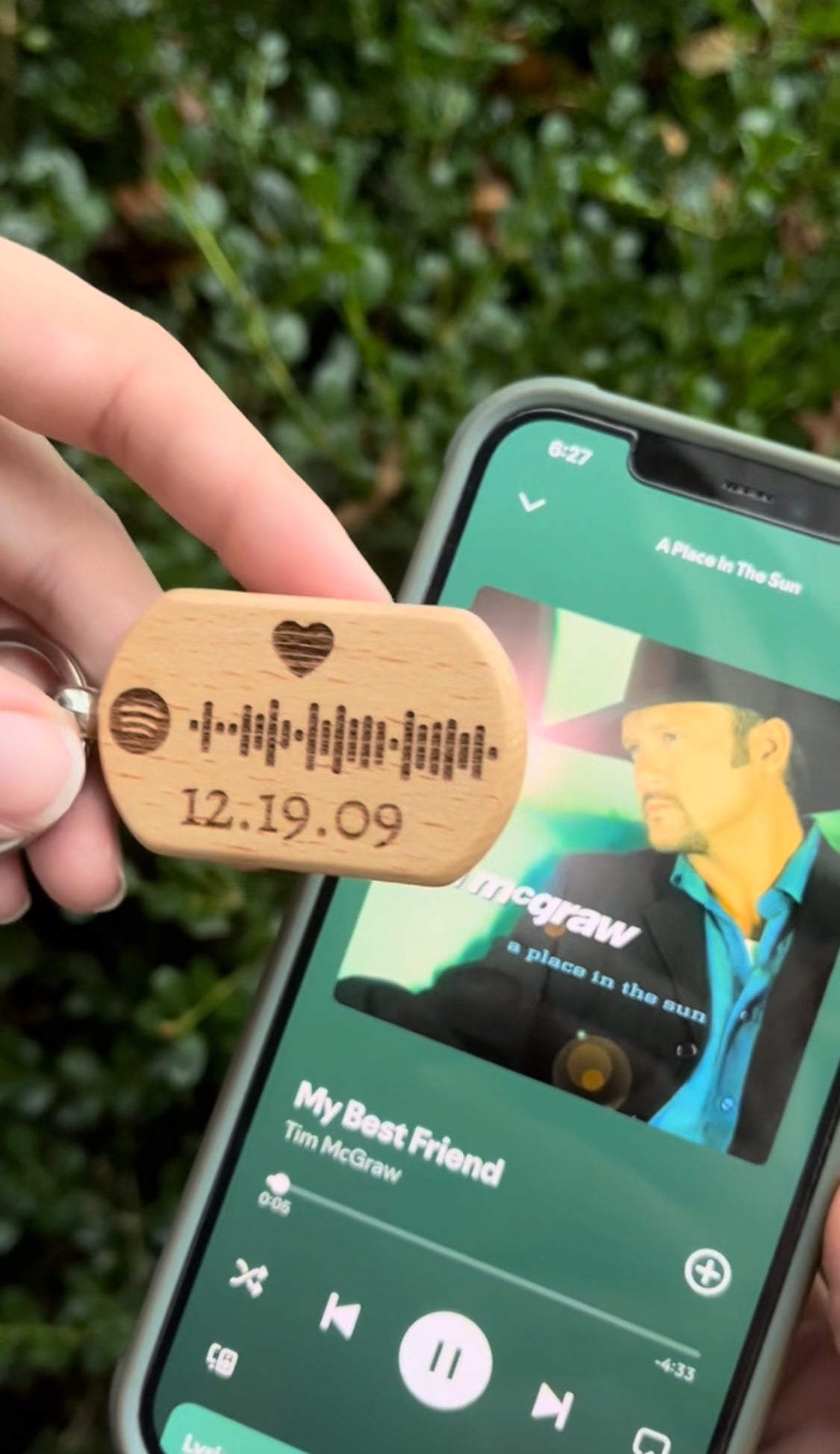 Wooden keychain with engraved date held in front of a smartphone playing music.