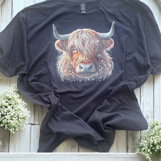 Black t-shirt with a highland cow design on a wooden surface with flowers.