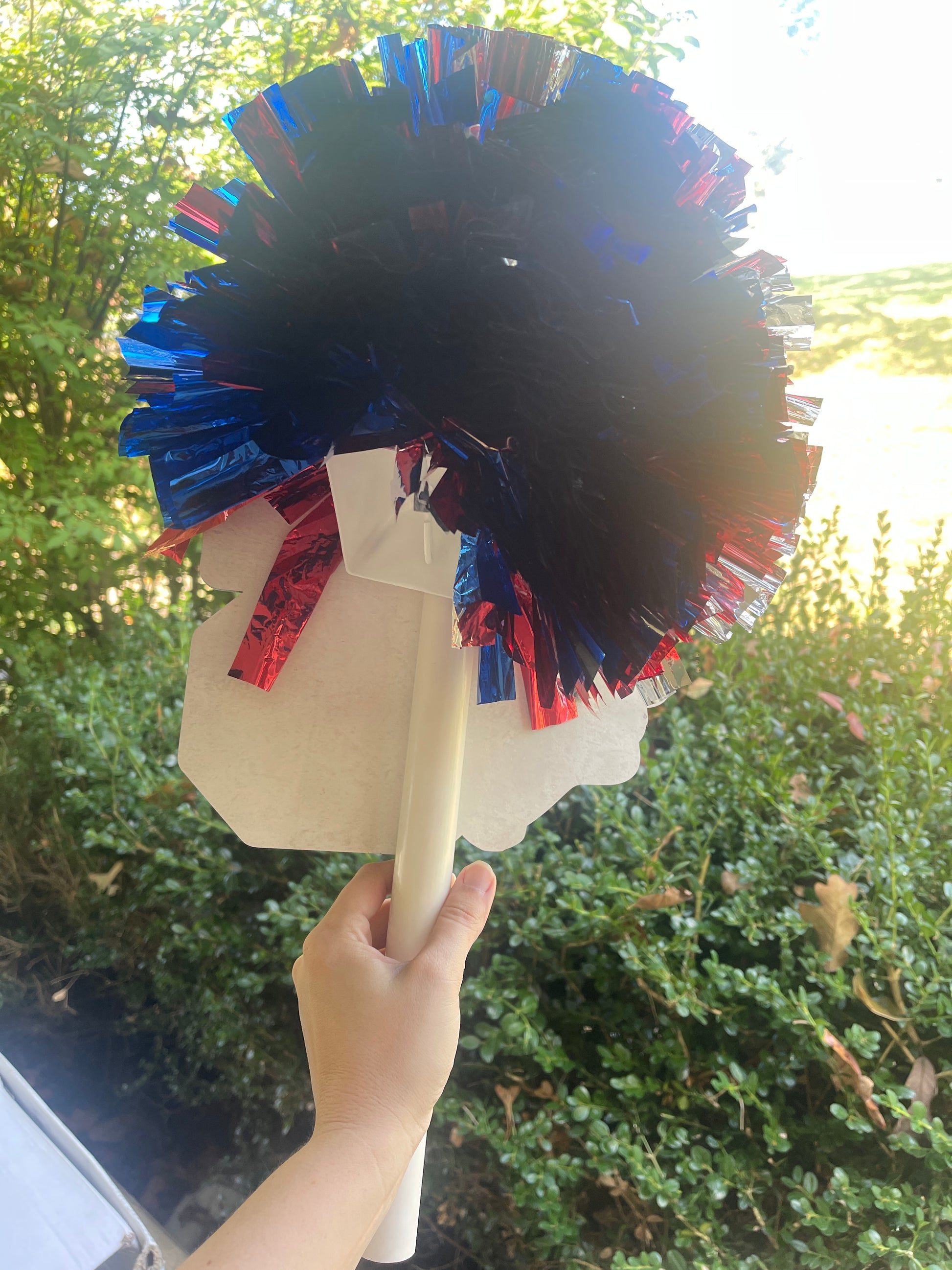 Hand holding a decorative fan with red and blue tassels against a blurred green outdoor background