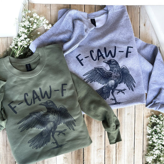 Two sweatshirts with a crow design and 'F-CAW-F' text on a wooden surface.
