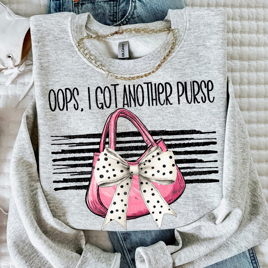 Gray sweatshirt with a graphic of a pink purse with a polka dot bow and text 'Oops, I got another purse' on a textured surface.