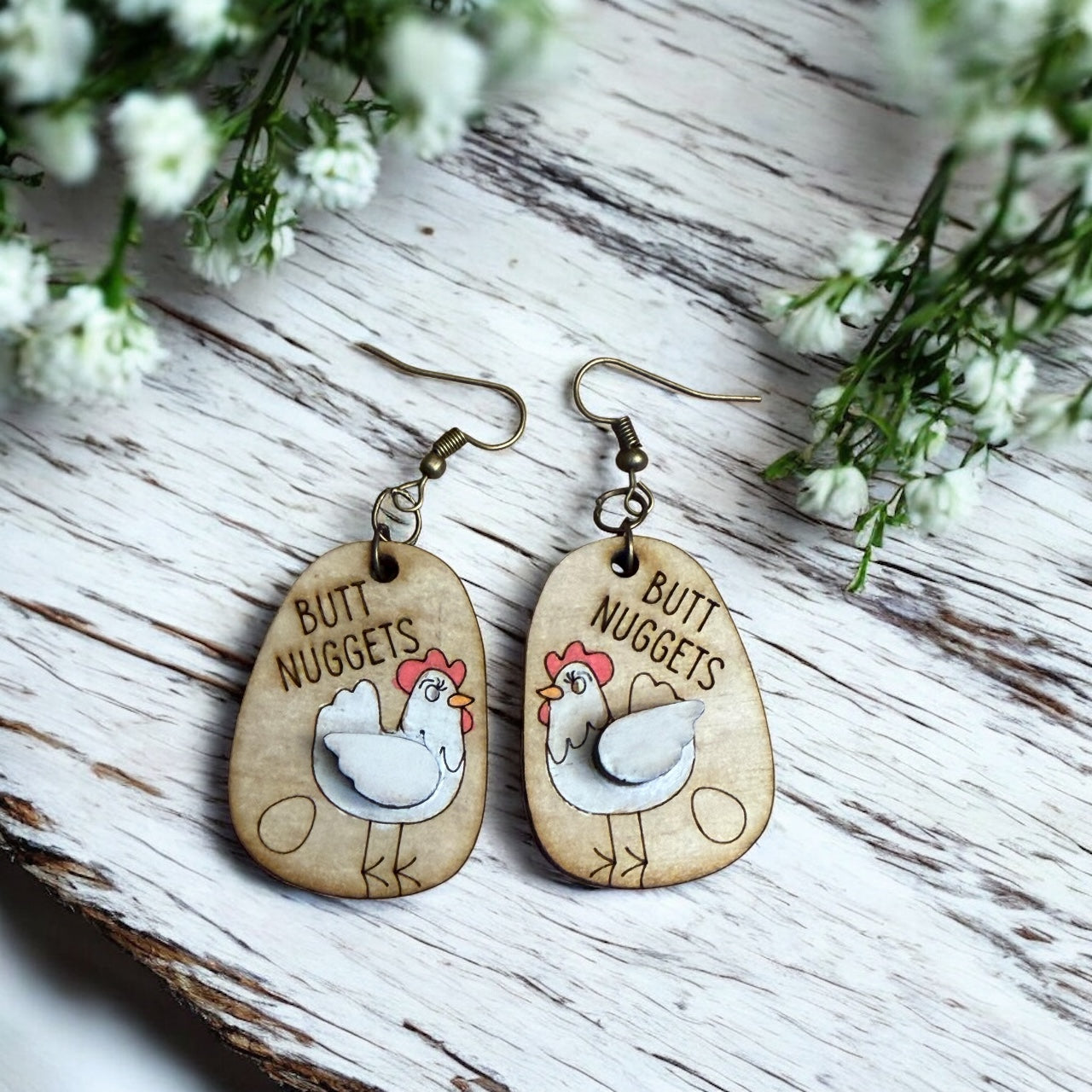 Wooden earrings with chicken design and 'Butt Nuggets' text on a wooden surface with flowers.