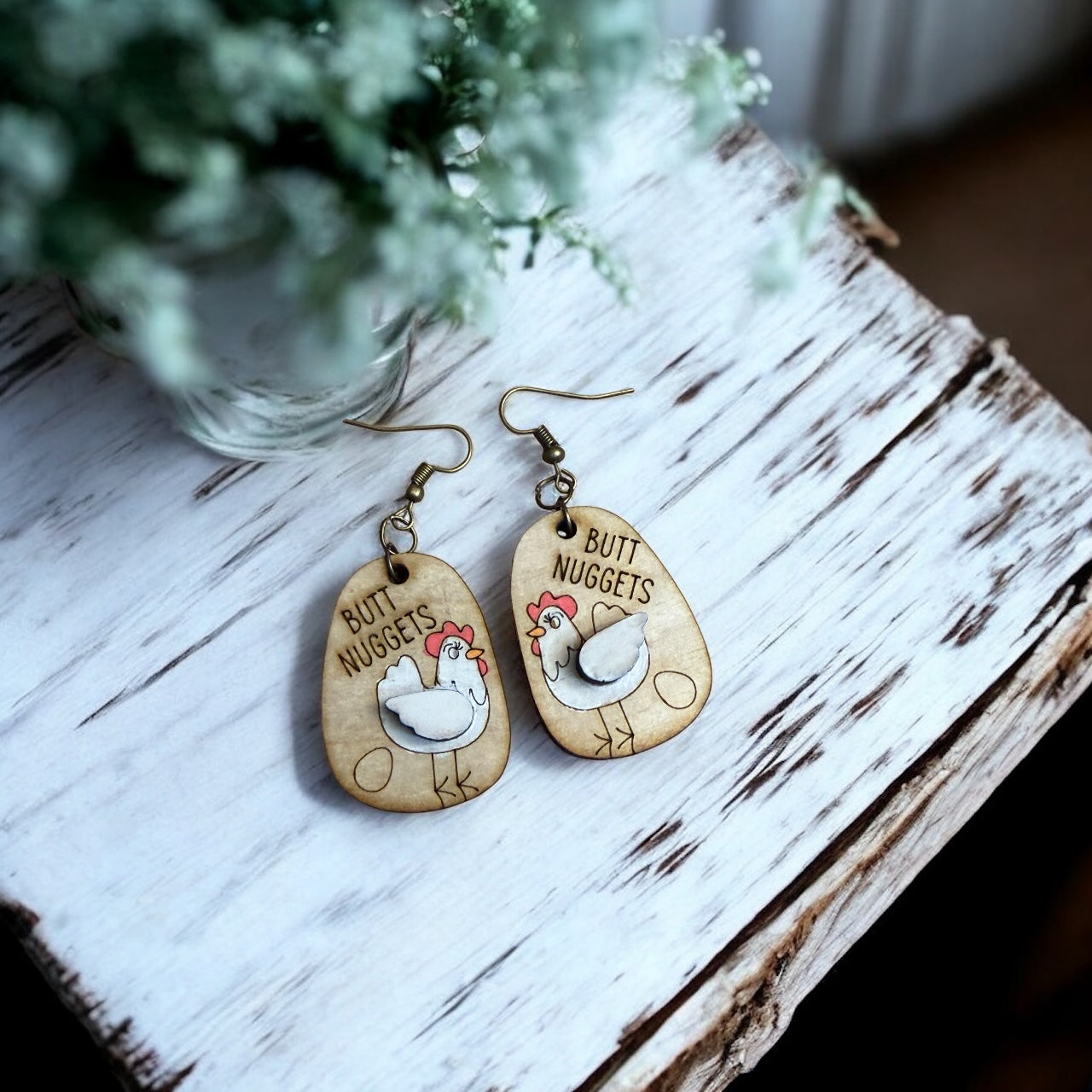 Wooden earrings with chicken illustrations and 'Butt Nuggets' text on a wooden surface.