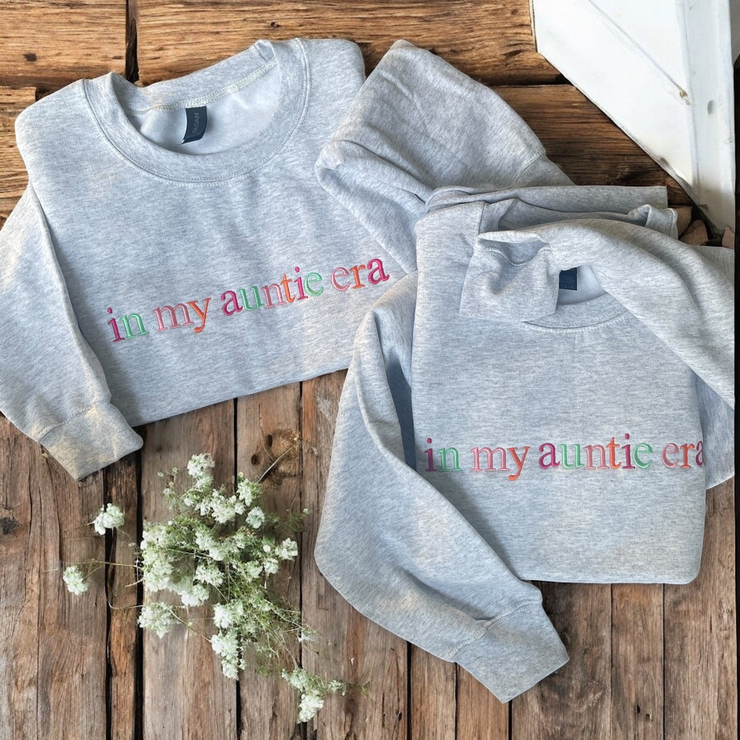 Two gray sweatshirts with 'in my auntie era' text on a wooden surface with white flowers.
