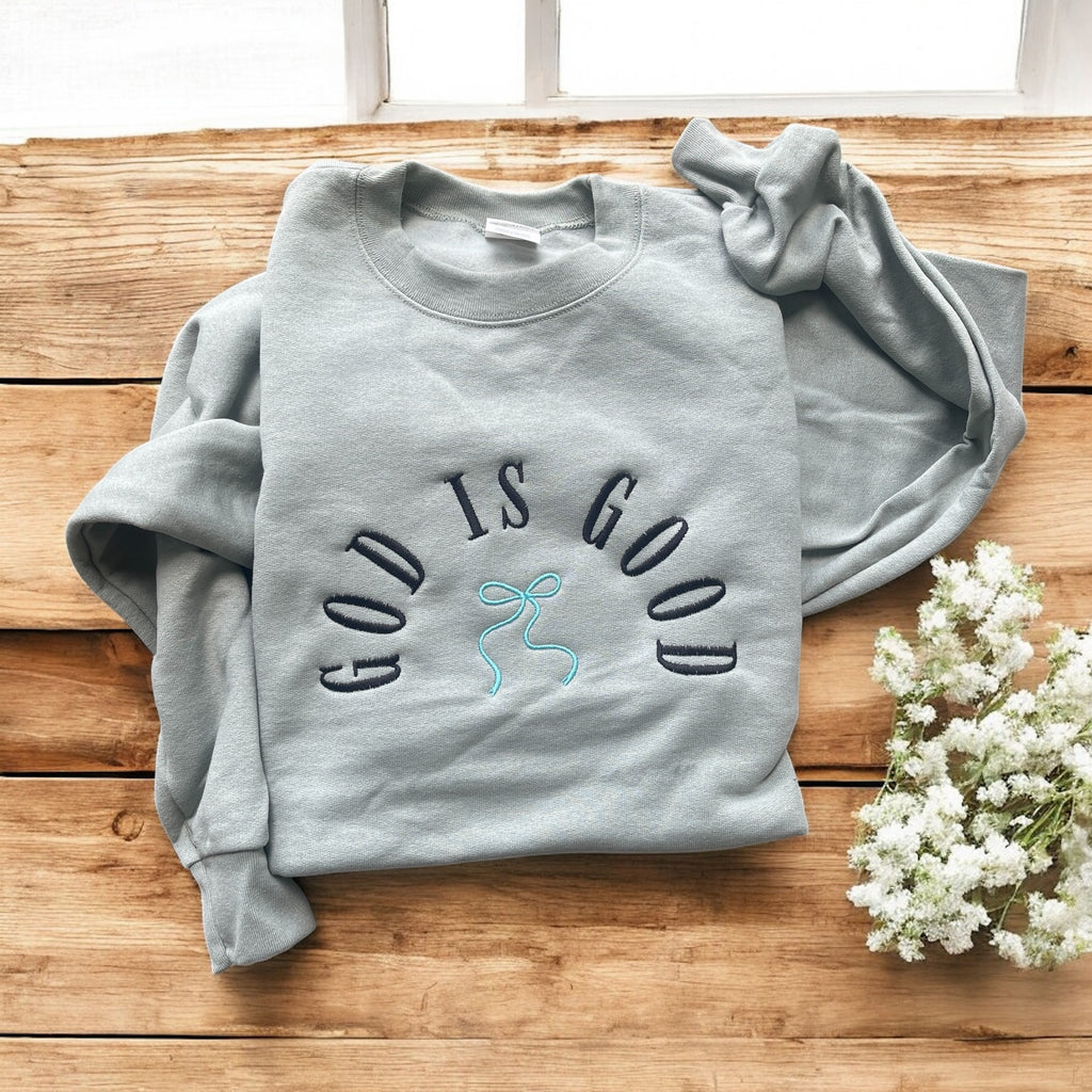 Gray sweatshirt with 'GOD IS GOOD' text on a wooden surface with flowers.