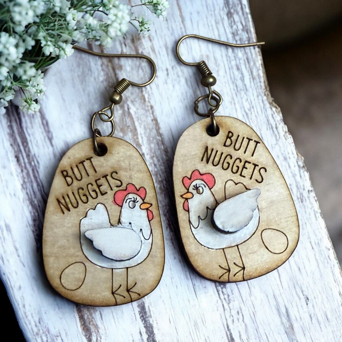 Wooden earrings with chicken illustrations and 'Butt Nuggets' text on a wooden surface.