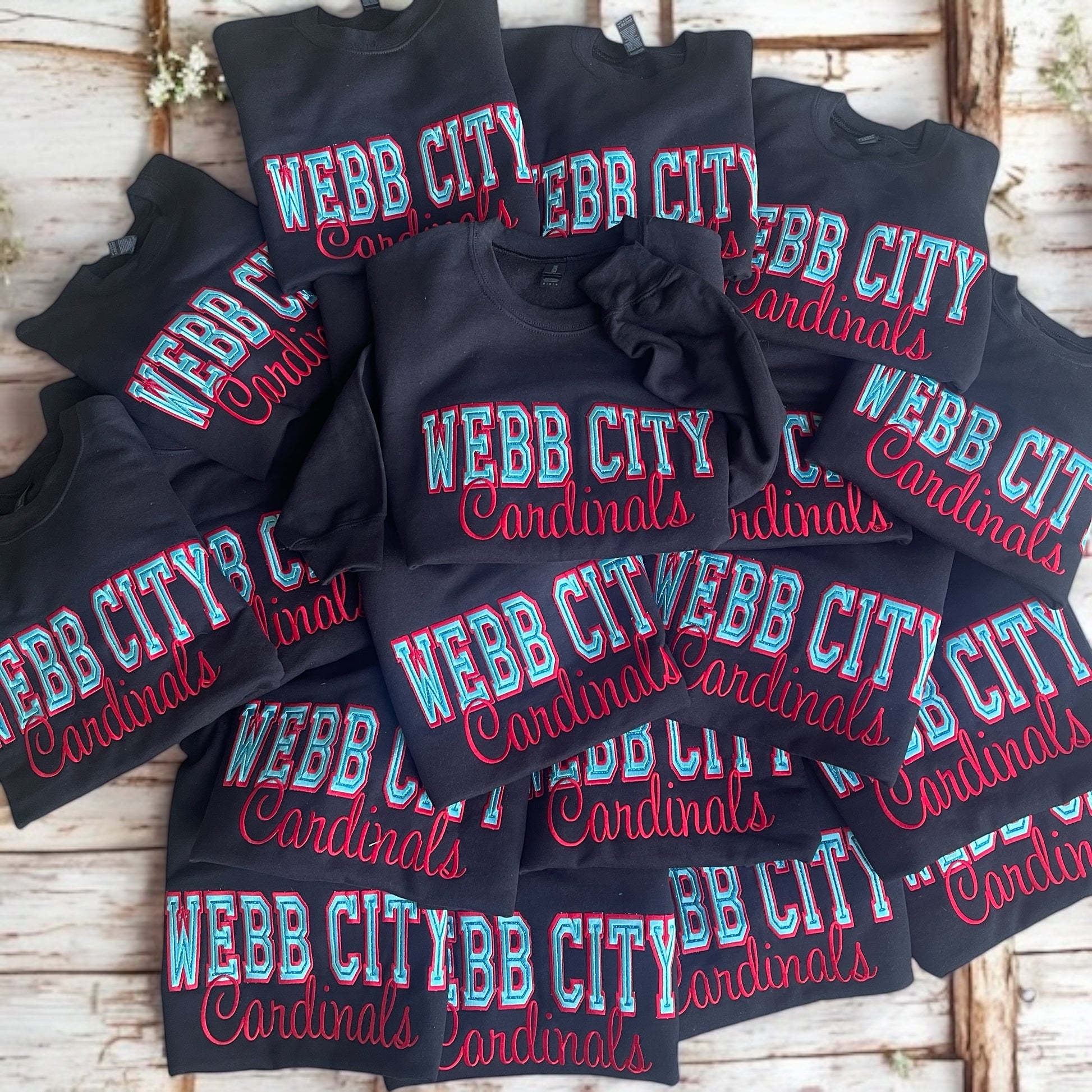 Stack of black t-shirts with 'WEBB CITY Cardinals' text on a wooden surface