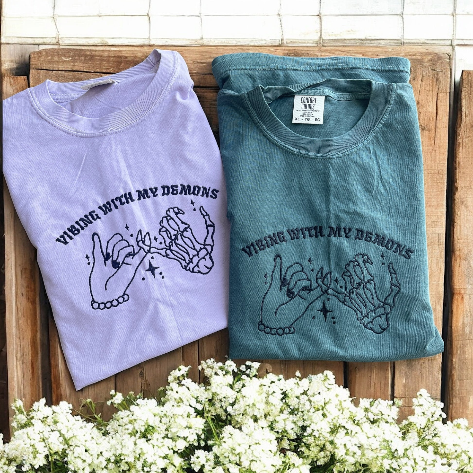Two folded t-shirts with a graphic design and text on a wooden surface with flowers in the foreground.