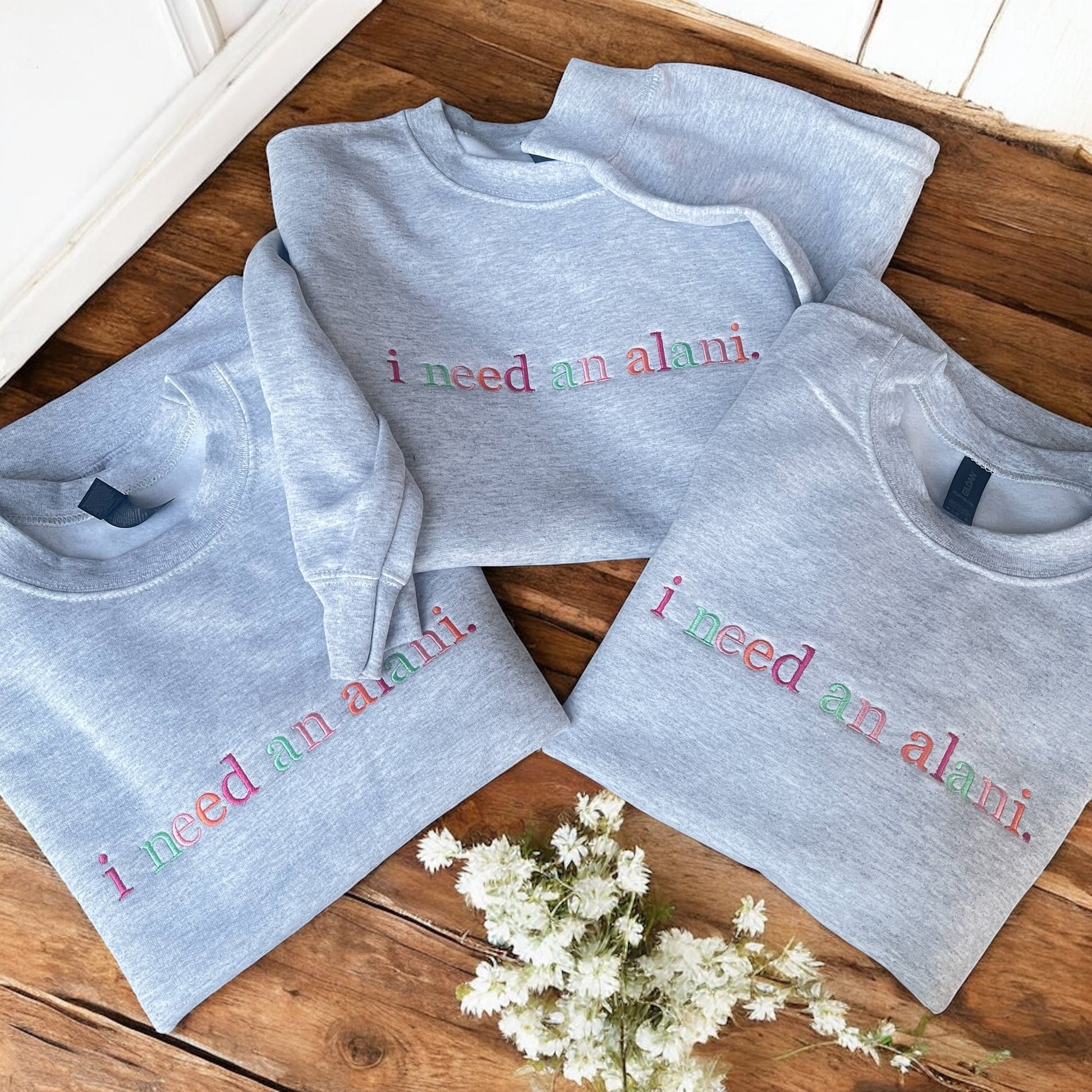 Gray sweatshirts with 'i need an alami' text on a wooden surface with white flowers.