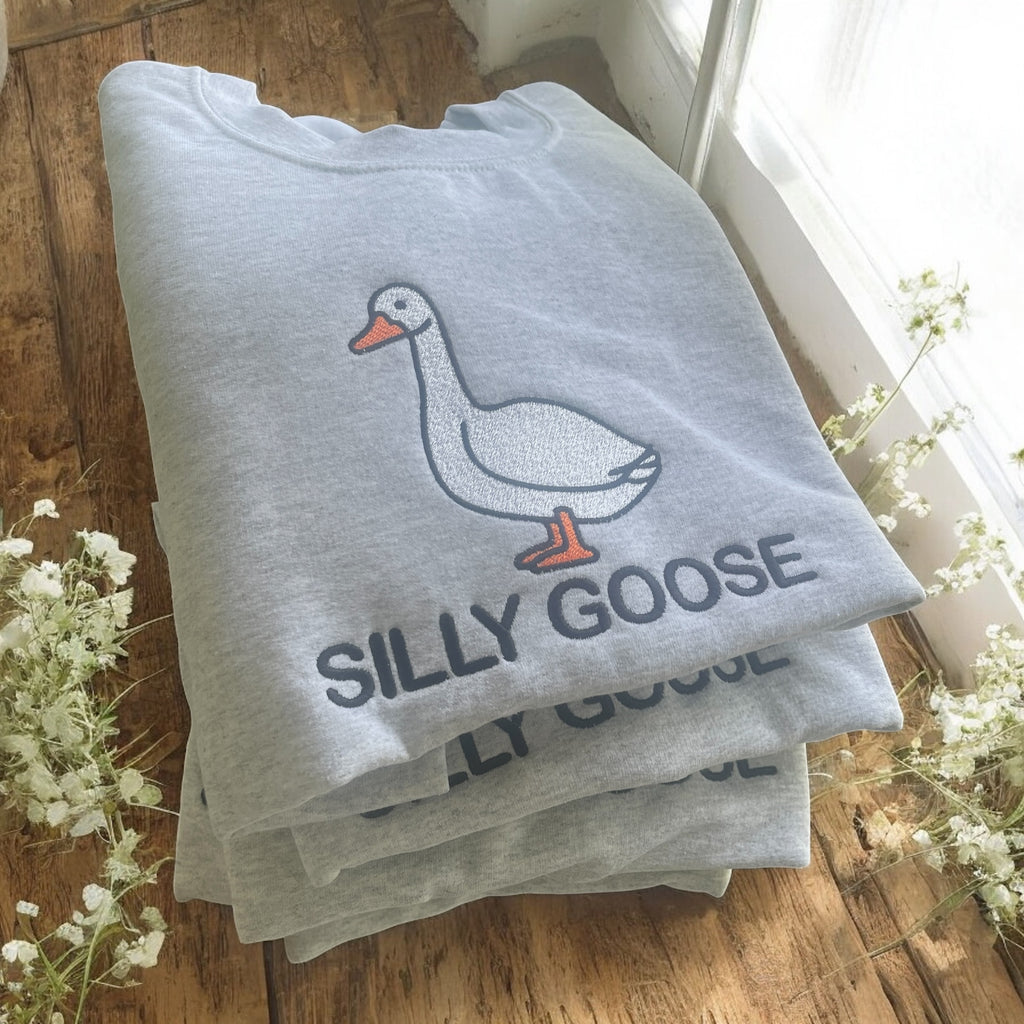 Stack of gray t-shirts with 'Silly Goose' design on a wooden surface with flowers.