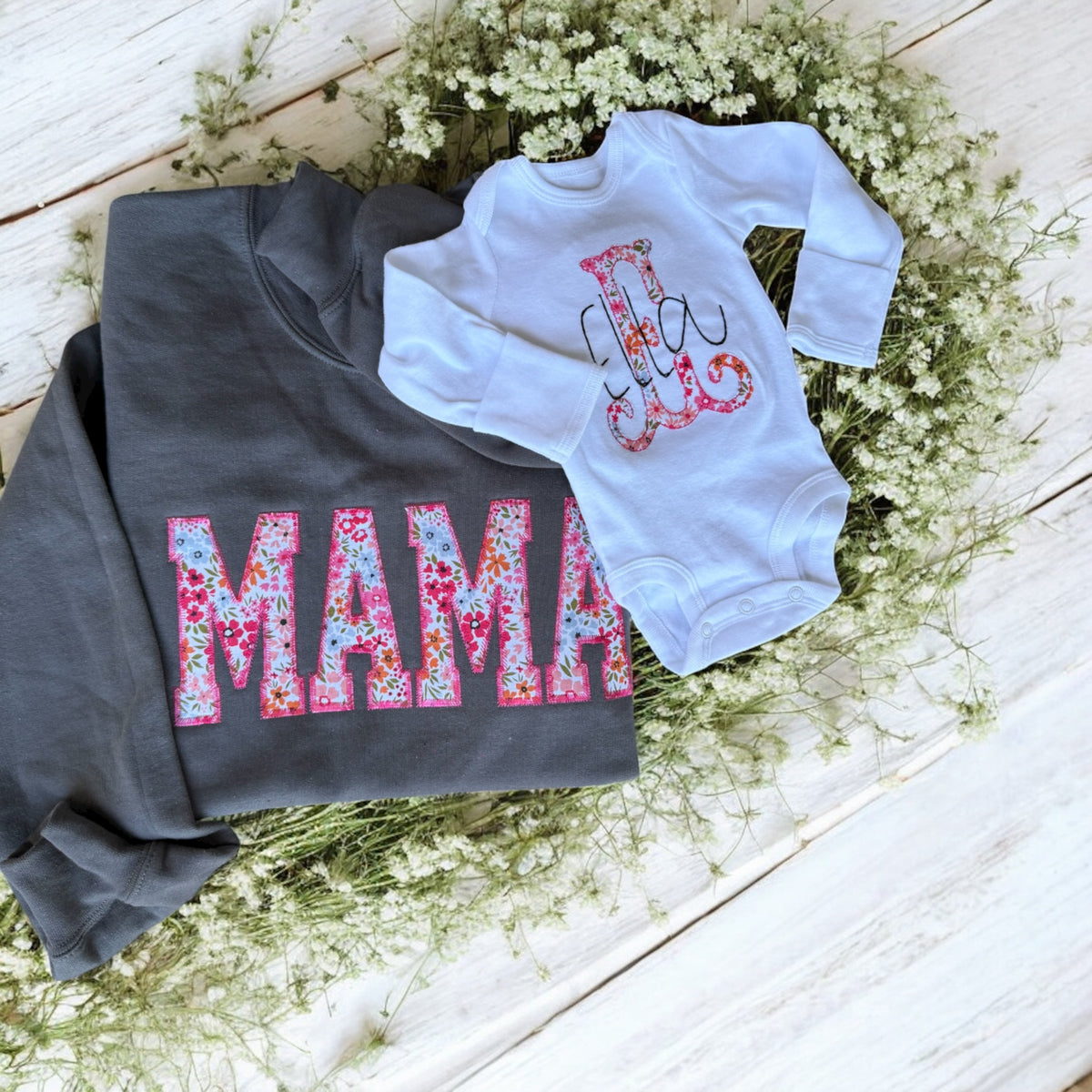 Gray sweatshirt with floral 'MAMA' text and white baby onesie with hand design on a wooden surface with flowers.