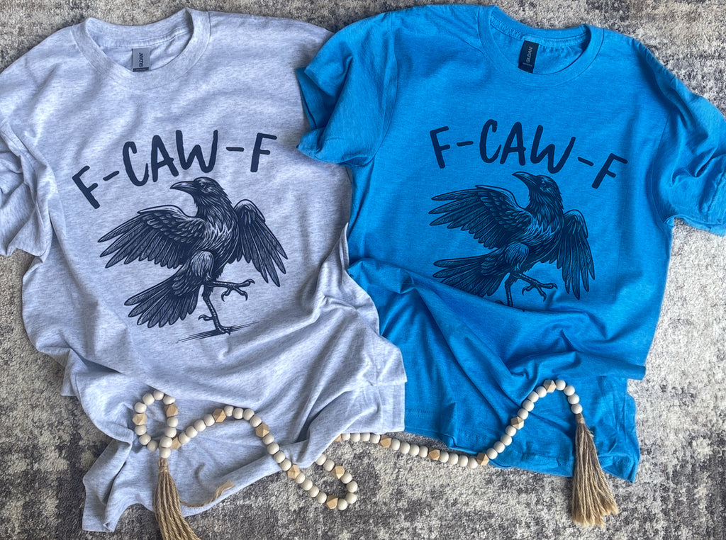 Gray and blue t-shirts with a crow graphic and 'F-CAW-F' text on a textured surface.