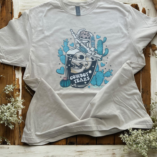 White t-shirt with a graphic design of a skull and cacti on a wooden surface with flowers.