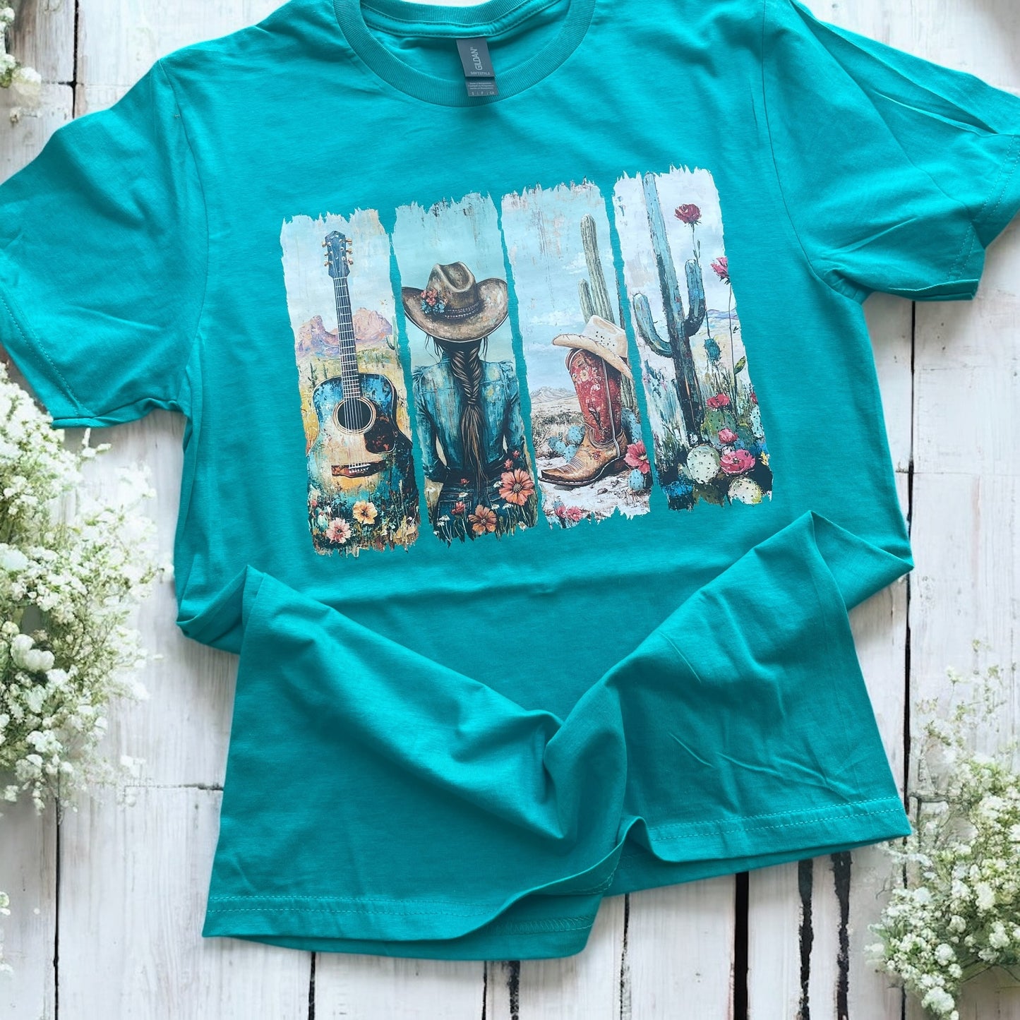 Teal t-shirt with a graphic design of a cowboy, guitar, and cactus on a wooden surface with flowers.