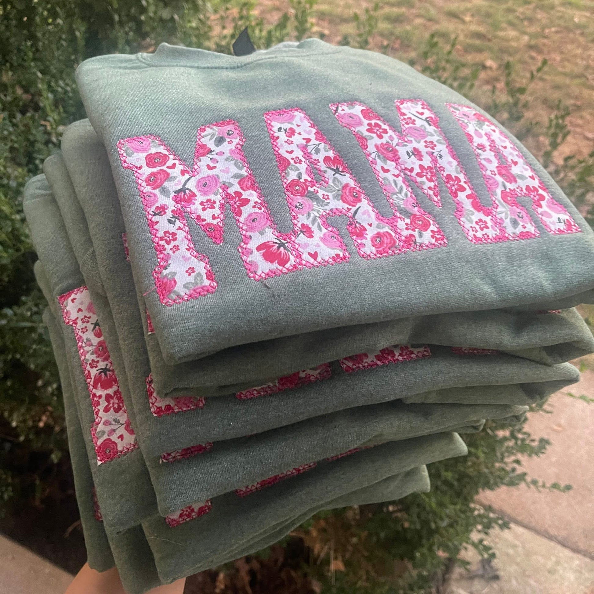 Stack of folded sweatshirts with floral 'MAMA' text in a garden setting