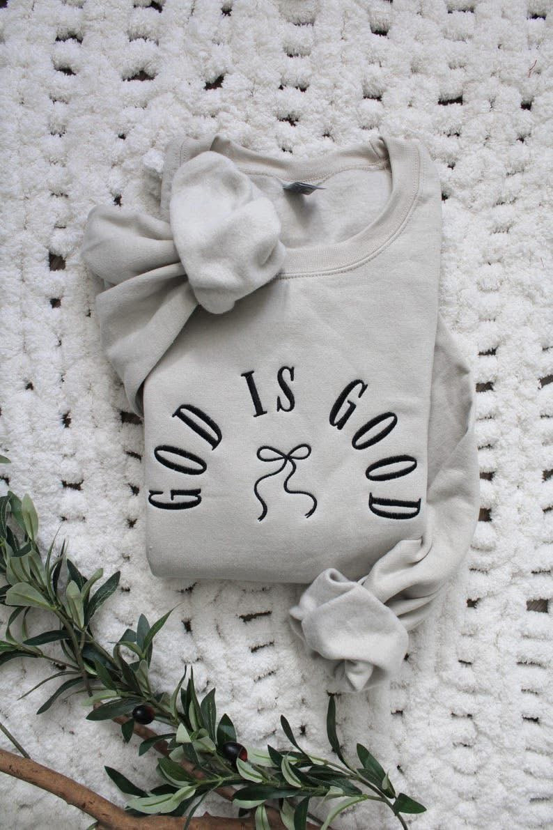 Light gray baby outfit with 'God is Good' text on a textured white surface with greenery.
