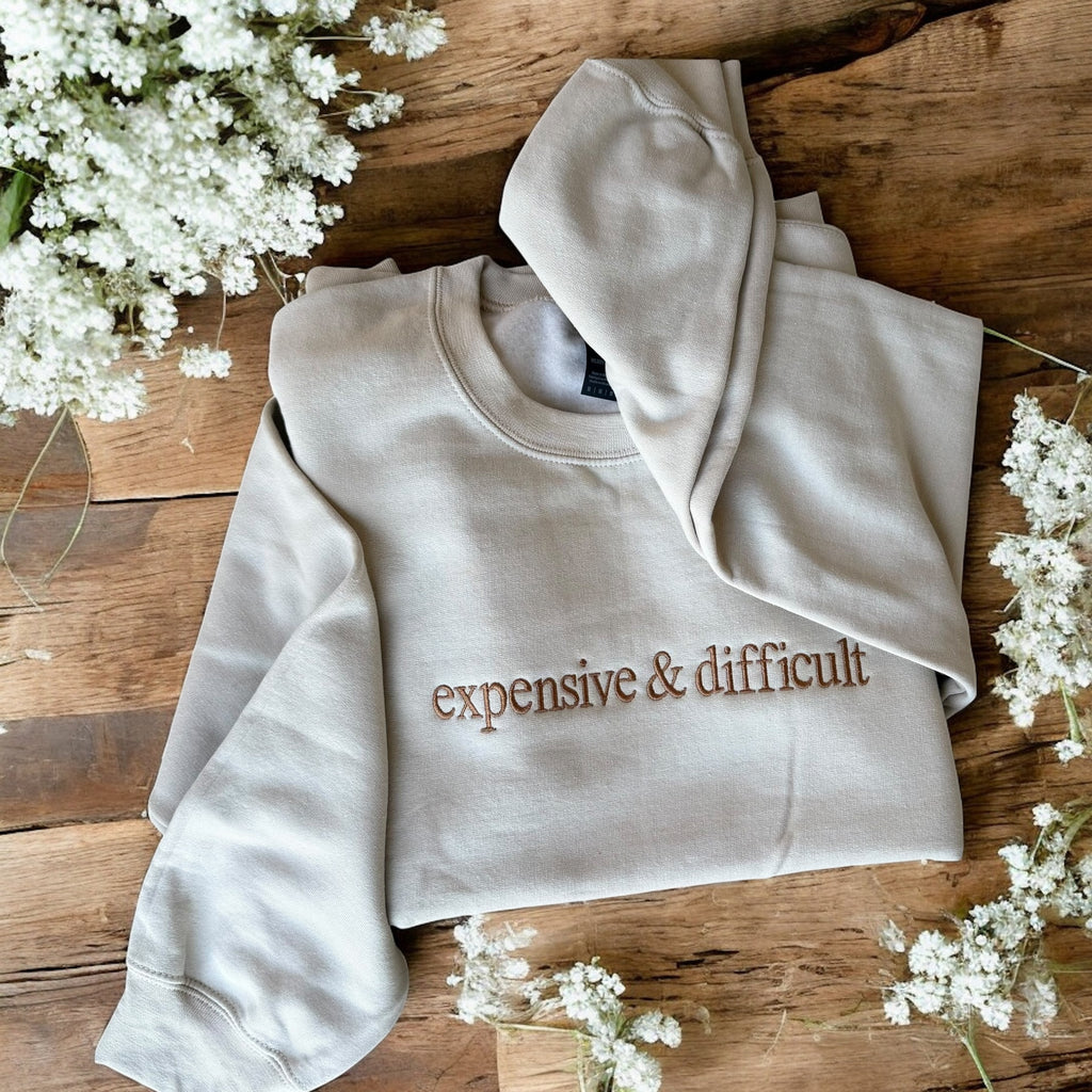 Beige hoodie with 'expensive & difficult' text on a wooden surface with white flowers.