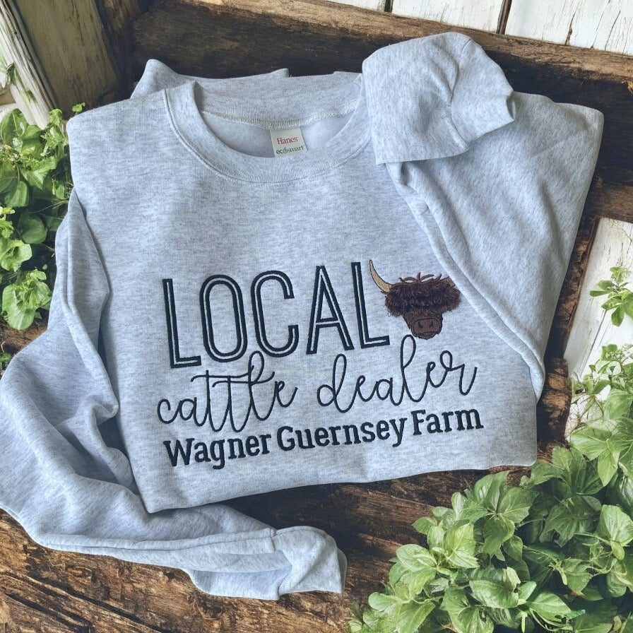 Gray sweatshirt with 'Local cattle dealer Wagner Guernsey Farm' text and logo on a wooden surface with plants.