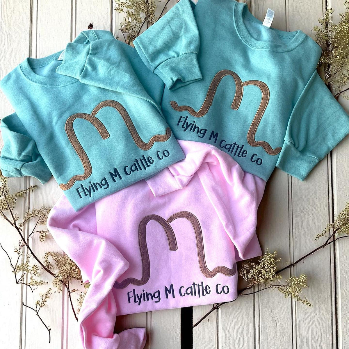 Two blue sweatshirts and a pink shirt with 'Flying M Cattle Co' branding on a wooden surface.