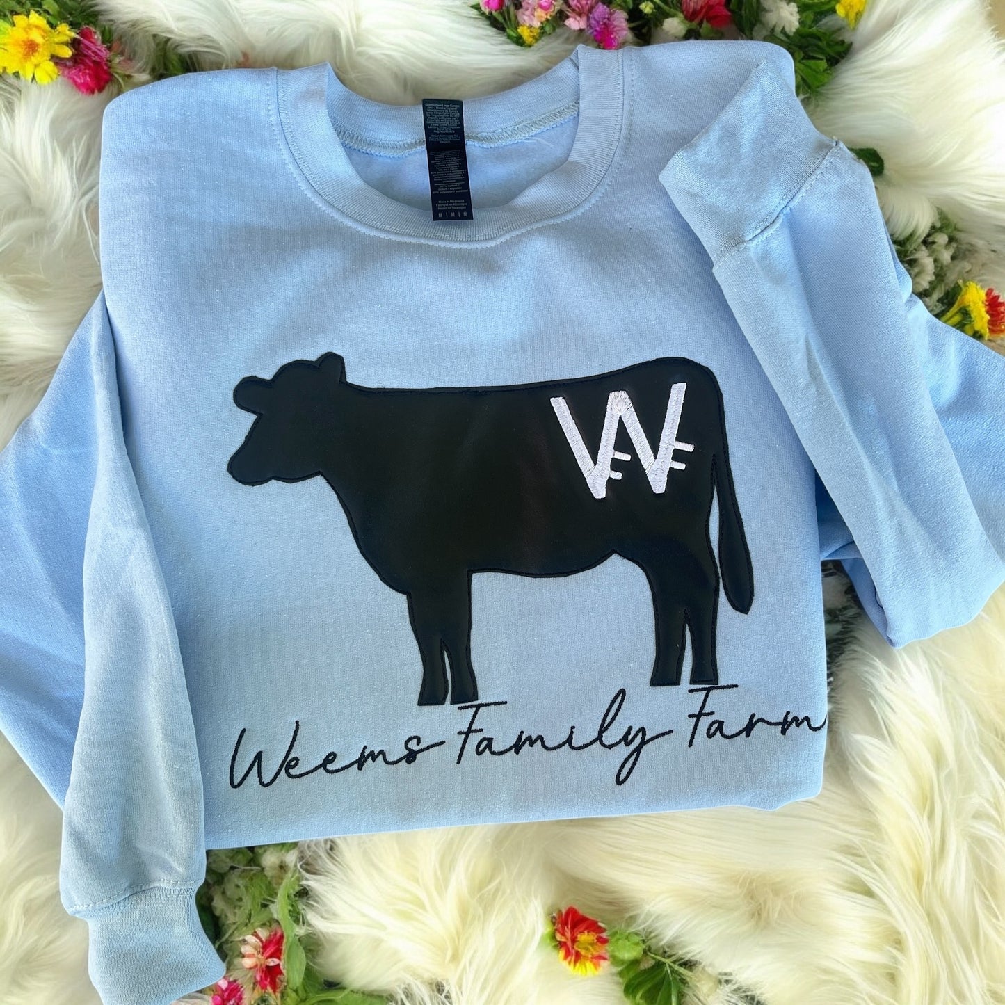 Light blue sweatshirt with a black cow graphic and 'Weems Family Farm' text on a fluffy white surface with flowers.