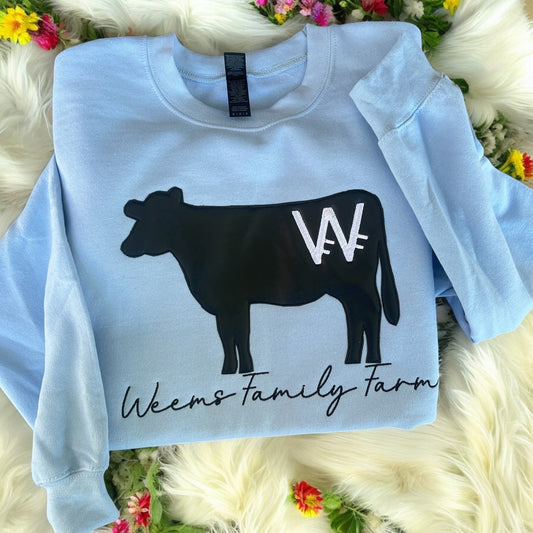 Light blue sweatshirt with a black cow graphic and 'Weems Family Farm' text on a fluffy white surface with flowers.
