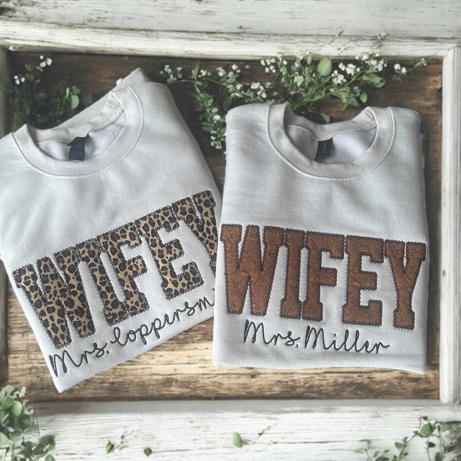 Two folded sweatshirts with 'WIFEY' text on a wooden surface with greenery.