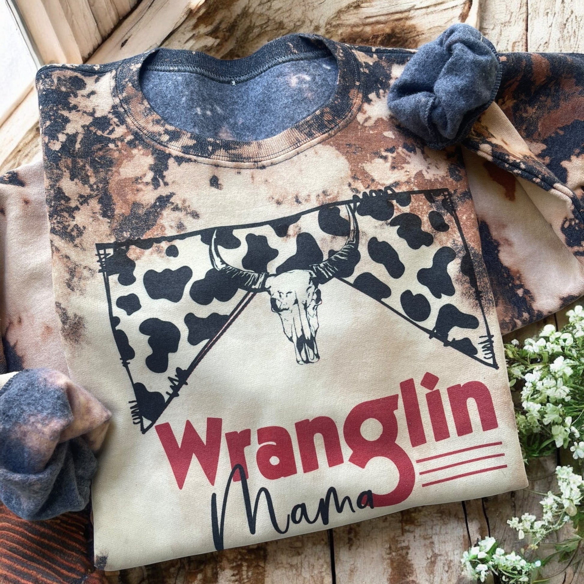 Sweatshirt with 'Wranglin Mama' design on a wooden surface with flowers.