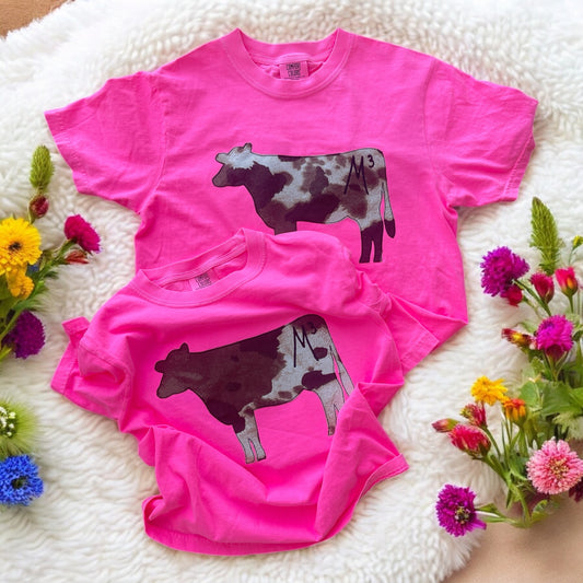 Two bright pink t-shirts with cow graphics on a white surface with flowers around.
