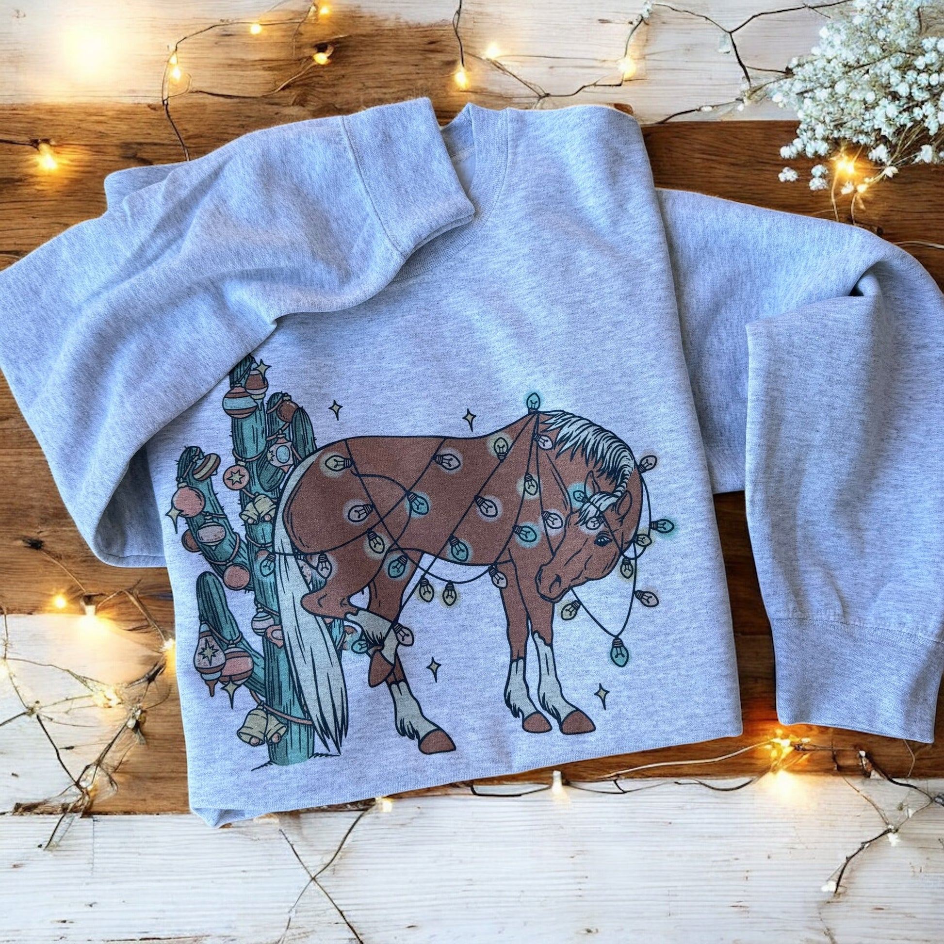 Gray sweatshirt with a horse and cactus design on a wooden surface with string lights.