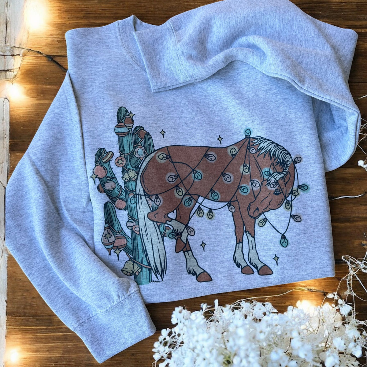 Gray sweatshirt with a horse and person design on a wooden surface with white flowers.
