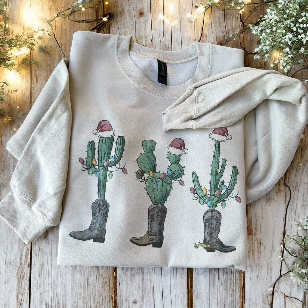 White sweatshirt with Christmas-themed cactus design on a wooden surface with decorative lights and plants.