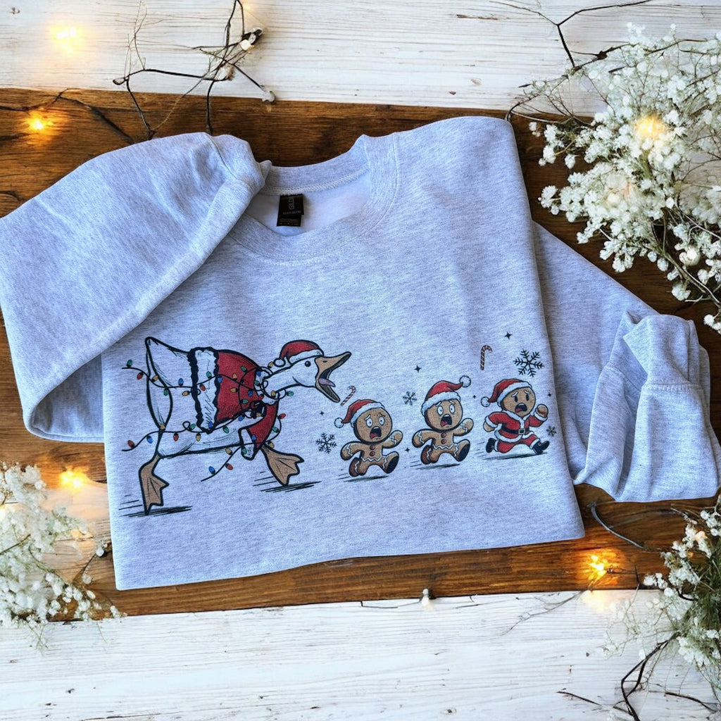 Gray sweatshirt with Christmas-themed design on a wooden surface with decorative lights and flowers.