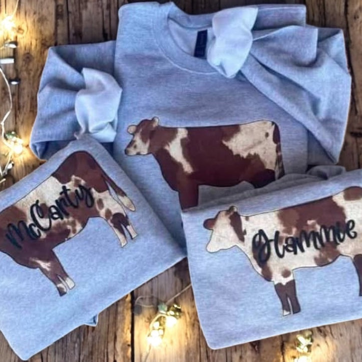 Gray sweatshirts with cow designs and names on a wooden surface with string lights.